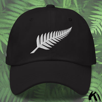 Black cap with white fern leaf design on a green leafy background
